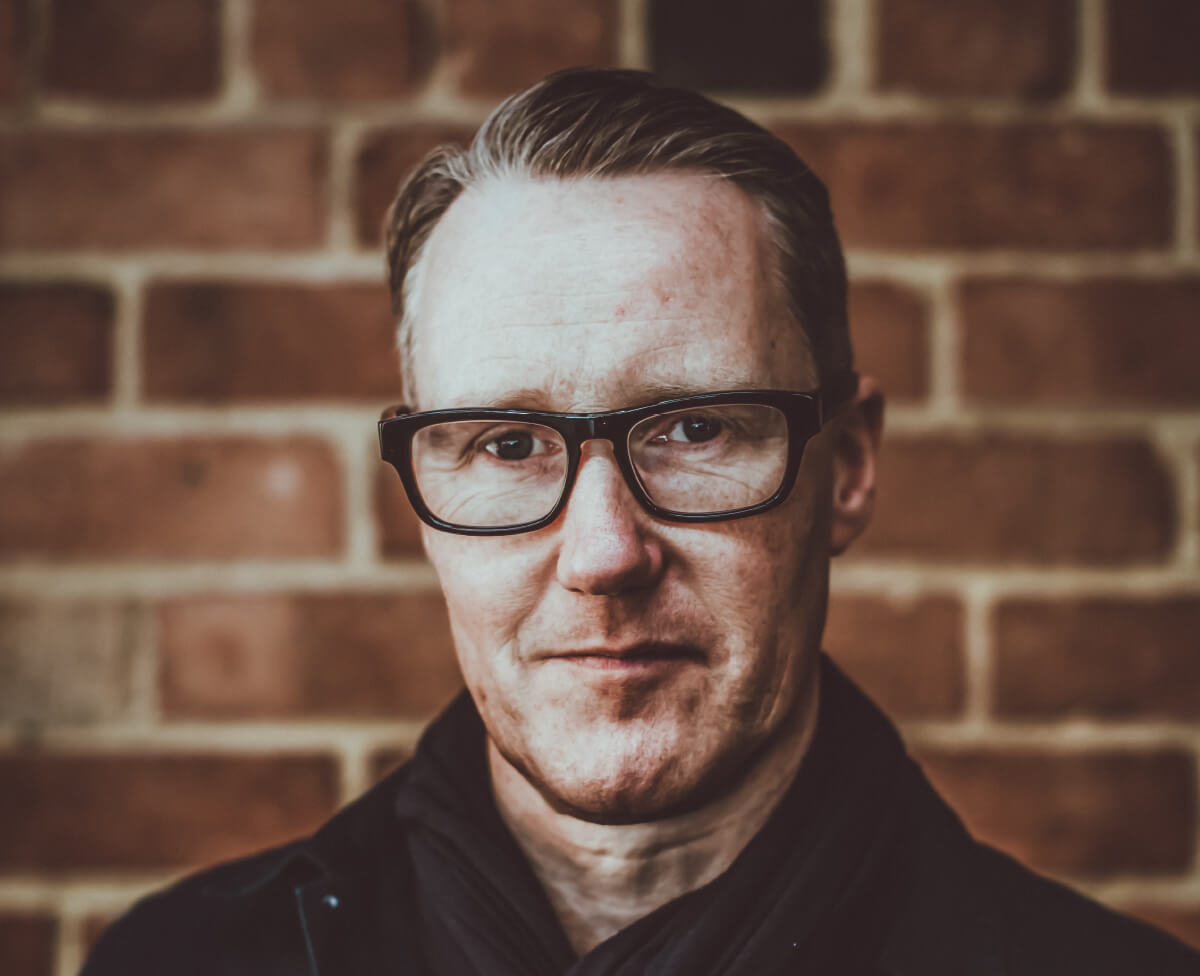 Portrait of Paul Paul Meikle-Janney, owner of Dark Woods Roastery, in front of brick wall.